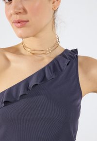 Morska off-shoulder majica s ruffled rubom, izrađena od teksturirane tkanine. Ima glatki izrez i slojevitu zlatnu ogrlicu.