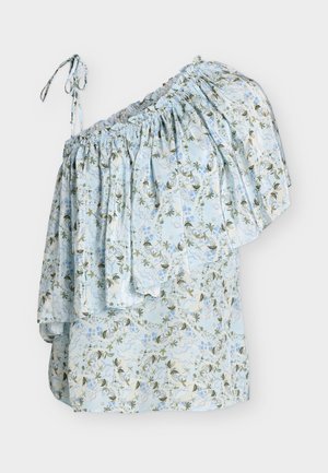 Femme portant un top asymétrique à bretelle unique bleu clair à fleurs avec volants, un jean large bleu foncé, des sandales blanches et un sac à main blanc.
