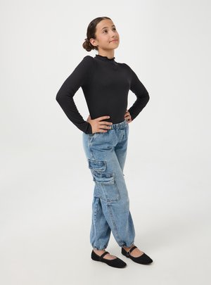 Ragazza giovane in piedi con sicurezza, mani sui fianchi, indossa una maglia nera a maniche lunghe, jeans cargo blu e scarpe Mary Jane nere su sfondo bianco.