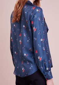 Niebieska bluzka w kwiaty z długim rękawem, zdobiona czerwonymi i białymi kwiatami. Wykonana z gładkiego materiału, o luźnym kroju i zapięciem na guziki przy mankietach.