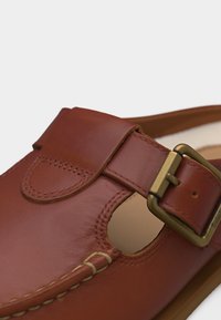 Sandale en cuir marron avec une grande boucle en laiton et un détail découpé ouvert sur le côté, montrant une couture lisse et une semelle intérieure souple.