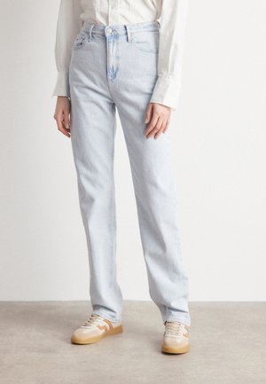 Persoon die lichtblauwe jeans met rechte pijpen, een wit overhemd met knopen en beige sneakers met rubberzolen draagt en op een eenvoudige vloer staat.