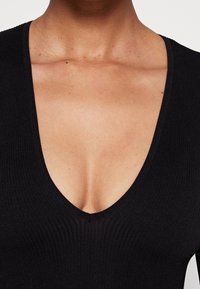 Nära bild av en persons övre bröst som bär en svart ribbad djup V-ringad klädnad. Hudtonen är medium med synliga fräknar och födelsemärken.