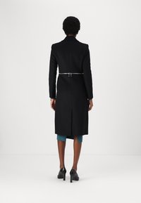 Cappotto lungo nero con silhouette aderente, stretto in vita da una cintura argentata, presenta un tessuto liscio e uno spacco sul retro.