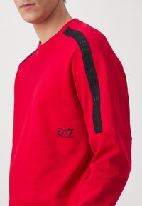 Sudadera roja con acentos negros en los hombros, que presenta el logo "EA7 Emporio Armani" en costura negra. Tejido suave con escote de cuello redondo.