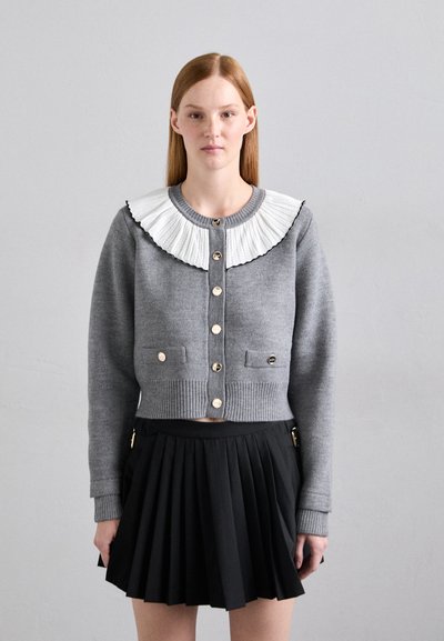 Maglione grigio corto con un colletto plissettato bianco, dettagli con bottoni dorati e due tasche frontali, abbinato a una gonna plissettata nera.