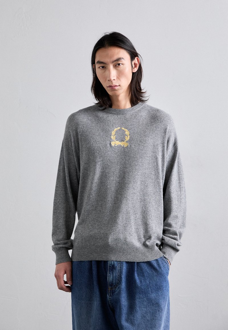 Grauer gestrickter Pullover mit langen Ärmeln, rundem Hals und gerippten Manschetten. Mit einem gelben gestickten Design auf der Brust. Lockere Passform.