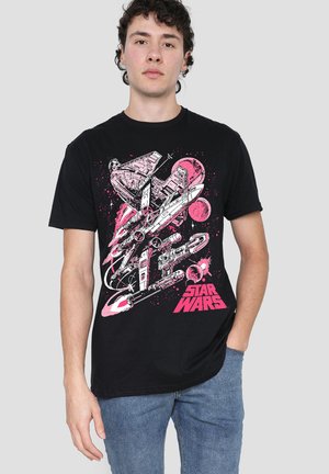 Junger Mann mit einem schwarzen Star Wars T-Shirt, das rosa und weiße Raumschiff- und Planetendesigns zeigt, kombiniert mit blauen Jeans vor einem grauen Hintergrund.