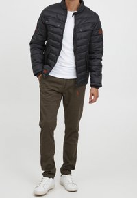 Veste matelassée noire avec fermeture éclair, deux poches poitrine et détails marron. Associée à un pantalon vert olive et des baskets blanches.