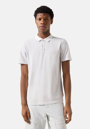 Young man standing facing forward, wearing a white short-sleeve polo shirt and white pants against a plain background.