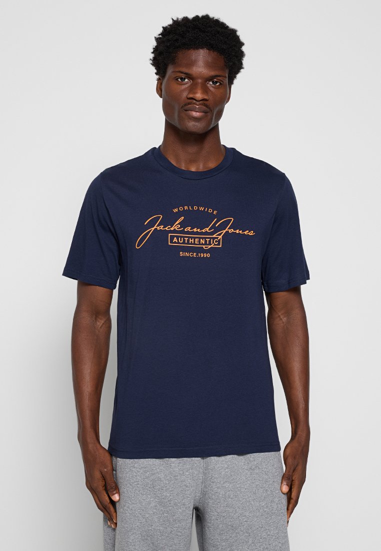Navyblaues Baumwoll-T-Shirt mit einem grafischen Druck in Orange. Kurze Ärmel, runder Ausschnitt und lockere Passform. Kombiniert mit grauen Shorts.