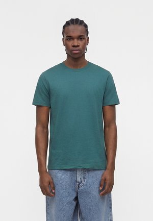 Joven con cabello trenzado, vestido con una camiseta verde lisa de manga corta y vaqueros de mezclilla azul claro, de pie frente a un fondo blanco.