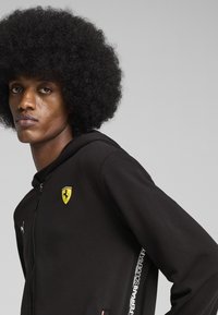 Sort zip-up hoodie lavet af blødt materiale, med et gult Ferrari-logo på brystet og en hvid stribe med "Ferrari Scuderia" på siden.