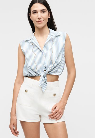 Γυναίκα που φοράει ένα αμάνικο, γαλάζιο και λευκό ριγέ κοντό τοπ δεμένο με διακοσμητικά από πέρλες και λευκά σορτς με υψηλή μέση και τσέπες με κουμπιά.