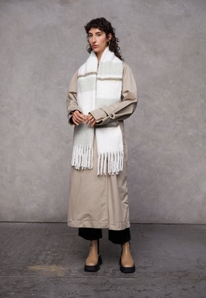 Beige trench coat with a textured finish, paired with a white scarf featuring green stripes and fringed edges. Black pants and beige ankle boots.