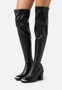 Stivali neri fino al ginocchio in pelle sintetica lucida, con tacco a blocco e accentuazione metallica sulla punta; texture liscia e design aderente.