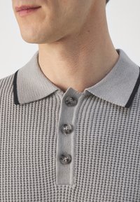 Polo gris con textura de waffle, con un ribete negro en el cuello y tres botones oscuros en la parte del cuello. Vista en primer plano del cuello y la parte superior del pecho.