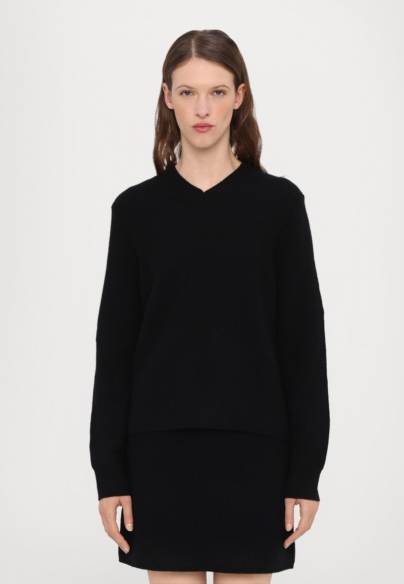 Maglione nero lavorato a maglia con scollo a V, maniche lunghe e vestibilità rilassata, abbinato a una gonna nera coordinata. Texture semplice, senza motivi o dettagli.