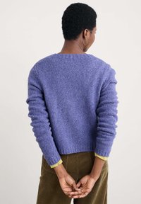 Person med kort, lockigt hår som bär en lila stickad tröja och bruna kordbyxor, stående med händerna knäppta bakom ryggen.