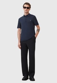 Marineblauwe poloshirt met een kraag, drie knopen en een klein logo. Draagt het met zwarte broeken en zwarte schoenen. Het model heeft een zonnebril op.