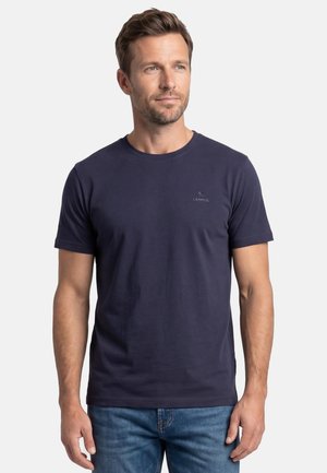 Man draagt een effen marineblauw T-shirt met korte mouwen en een blauwe spijkerbroek, staande tegen een witte achtergrond, iets schuin kijkend.