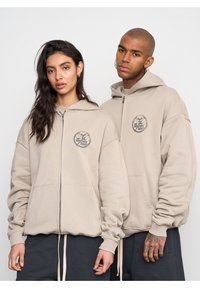 Sudaderas beige con cremallera fabricadas en un tejido suave, que presentan grandes bolsillos frontales y un logotipo impreso en el área del pecho izquierdo, combinadas con pantalones oscuros.