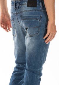 Un jean en denim bleu clair présente une coupe fuselée, des poches arrière avec un détail de couture et une étiquette en cuir noir sur la taille.