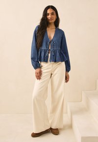 Plava i bijela bluza na kockice s dugim rukavima, V-izrezom, zatvaranjem na vezanje i strukom s peplum detaljem. Uparena s kremastim hlačama širokih nogavica i smeđim cipelama.