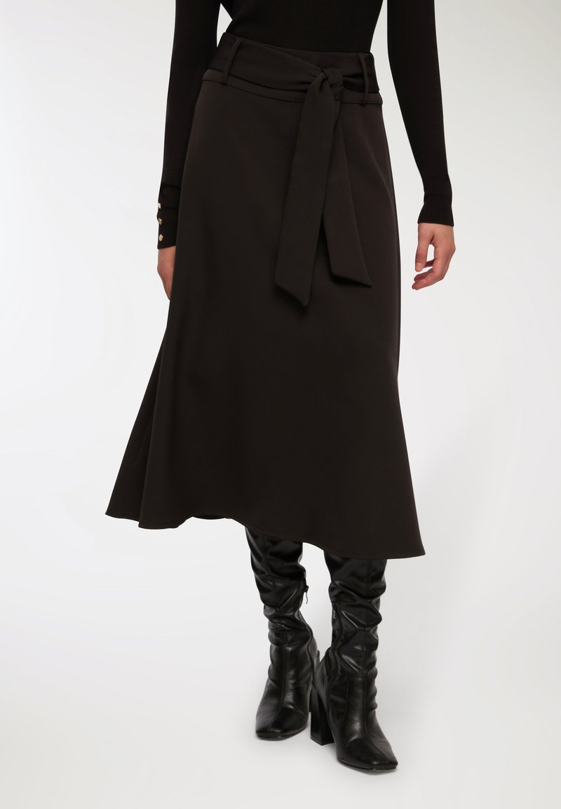 Gonna nera a trapezio con una cintura in tessuto legata in vita, materiale fluido e superficie liscia, abbinata a stivali neri fin sopra il ginocchio.