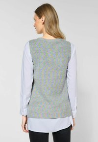 Gilet lavorato a maglia con una trama multicolore; le tonalità grigie, rosa e verdi creano un design a motivi. Indossato sopra una camicia a maniche lunghe bianca.