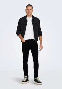 Juodas džinsinis švarkas dėvimas ant baltos marškinėlių, derinamas su juodais siaurais džinsais ir juodais sportbačiais su kremine padu, pateikti ant neutralaus fono.