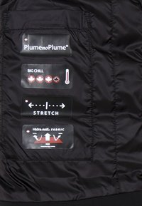 Giacca nera isolante in tessuto con numerose etichette che descrivono le caratteristiche: "Plume no Plume," "Big Chill," "Stretch," e "Hidra-nek Fabric."