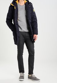 Parka marine isolée avec une capuche doublée de jaune, fermeture éclair à l'avant et patch logo. Portée avec un pull gris, un pantalon noir et des chaussures grises.