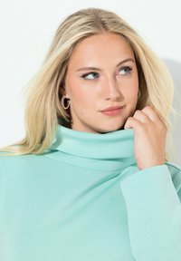 Miętowo zielony golf z gładkiego materiału, o luźnym kroju i długich rękawach. Dekolt jest złożony i luźno opadający, dodając szczegółu.