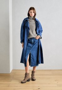 Manteau trench en denim avec large col, col roulé gris côtelé, jupe en denim avec fente avant, et bottes de cheville en cuir marron à lacets avec un talon bloc.