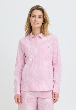 Mujer con cabello rubio hasta los hombros, vistiendo una camisa de manga larga con rayas verticales rosas y blancas y pantalones a juego, sobre un fondo blanco.