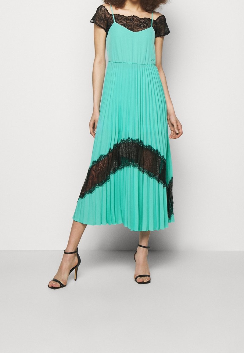Un vestido midi plisado de color verde azulado con detalles de encaje negro en el corpiño y el dobladillo, con tirantes finos y una silueta fluida. Sandalias negras de tacón.