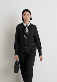 Blouse noire à manches longues avec un motif texturé, boutonnière à l'avant et col rond. Associée à un pantalon noir et un petit sac à main noir.