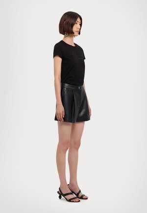 T-shirt nero in cotone con maniche corte e logo ricamato, abbinato a una minigonna plissettata in pelle nera e sandali neri con lacci.