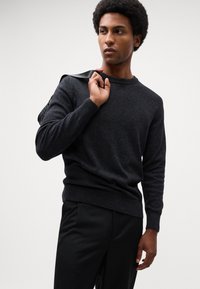 Dark gray knit sweater with a round neckline and ribbed cuffs, paired with black trousers. Model holds a textured bag over one shoulder.