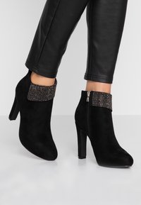 Botines de ante negros con tacón alto, que presentan un puño de tela negra con tachuelas y un cerradura lateral con cremallera.