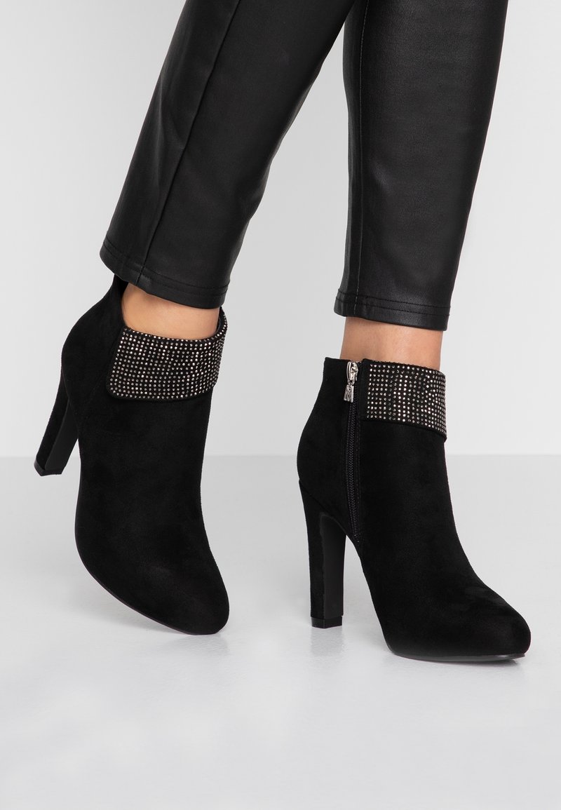 Botines de ante negros con tacón alto, que presentan un puño de tela negra con tachuelas y un cerradura lateral con cremallera.