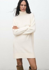 Abito turtleneck lavorato a maglia di colore crema, con maniche lunghe, polsini e orlo a coste. Il tessuto sembra morbido ed è lungo fino a metà coscia.