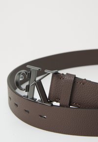 Bruine gestructureerde leren riem met een zwarte metalen gesp waarop de letters "cK" op een witte achtergrond zijn weergegeven.