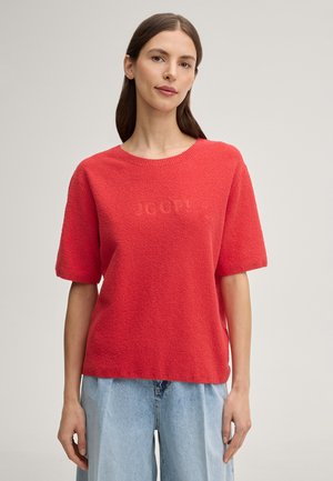 Kobieta w koralowym, krótko rękawowym swetrze o fakturze, zestawionym z jasnoniebieskimi, wysokim stanem jeansami, stojąca na jednolitym jasnym tle.