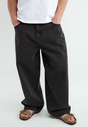 Osoba nosząca luźne czarne jeansy z rękami w kieszeniach, w połączeniu z cielistymi sandałami z odkrytymi palcami i białą koszulą, stojąca na jednolitym tle.