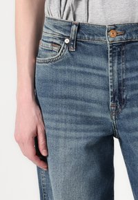 Světle modré džíny s vysokým pasem, s měděnými knoflíkovými detaily, klasickým pětipocket designem a jemným vyblednutím po nohách.