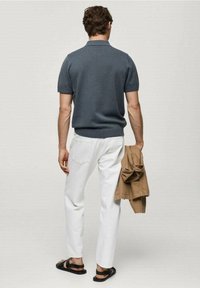 Harmaa lyhythihainen polo-paita, valkoiset slim-fit-housut, mustat sandaalit. Pitelee vaaleanruskeaa takkia, nähtynä takaa.