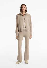 Beigefärgad stickad outfit bestående av en jacka med krage och figurnära utsvängda byxor, tillverkad av ett mjukt, texturerat tyg, i kombination med ljusa sneakers.