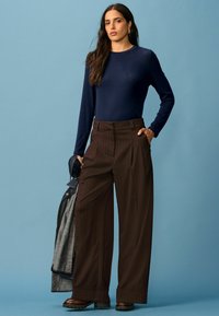 Mere pikkade varrukatega sinine särk, mis on ühendatud pruunide triibuliste laia säärega pükstega. Ühe käe peal on visatud teksajakk. Taust on helesinine.